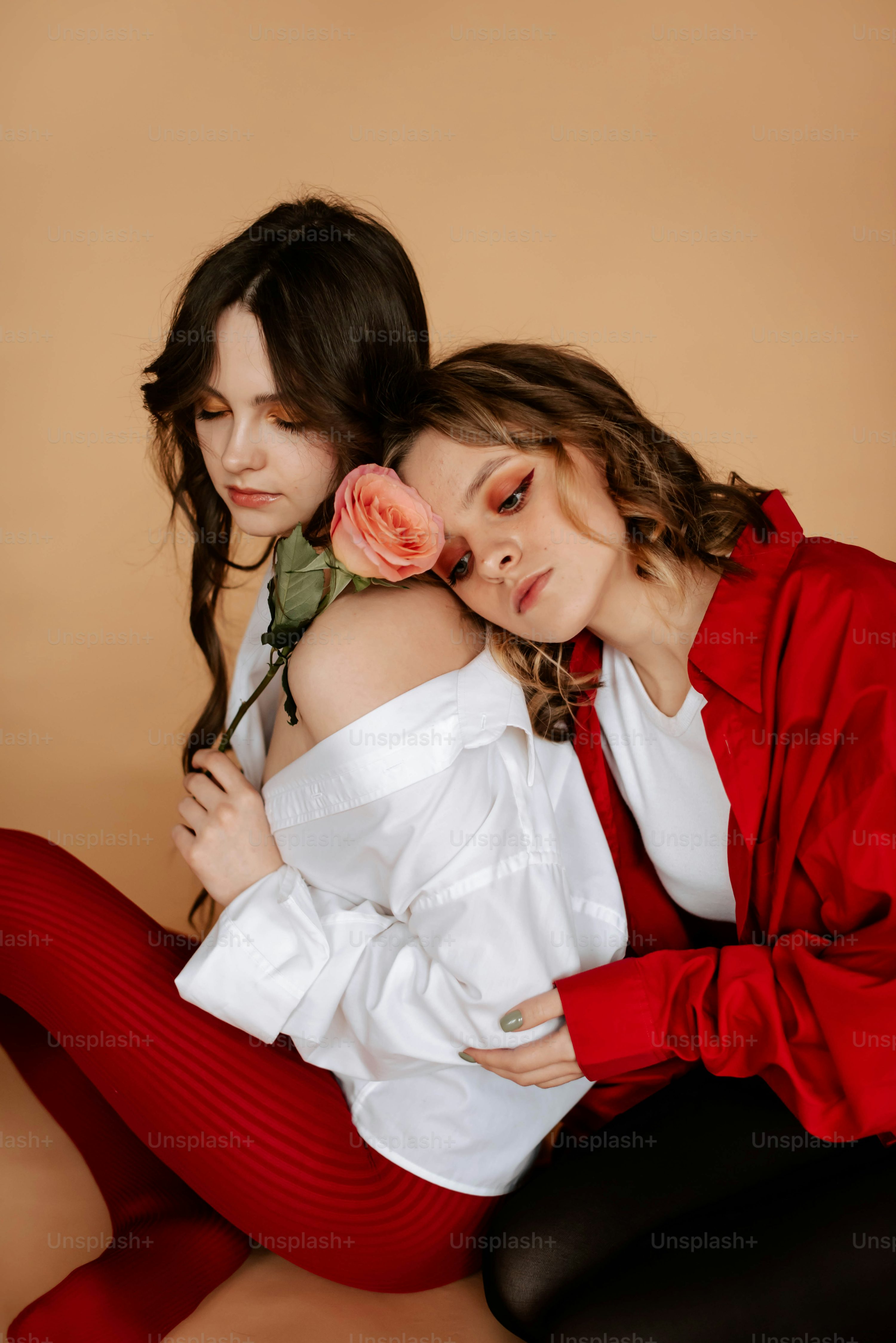 the image shows two young women embracing each other. one woman is wearing a white shirt and red ribbed leggings, while the other is wearing a red shirt and black leggings. they are sitting close together, with one woman's head resting on the other's shoulder. a single pink rose is held between them, adding to the tender and intimate feeling of the scene. the background is a muted beige, which keeps the focus on the women. the lighting is soft and even, creating a pleasant and calming atmosphere. the overall mood is one of friendship, affection, and closeness.