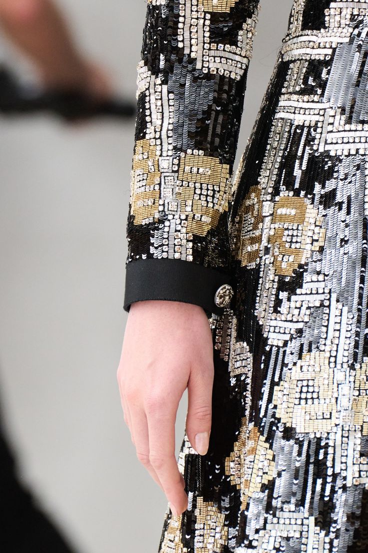 a closeup shot of a woman wearing a black and silver dress with intricate sequins and embroidery. the dress features a detailed design and a luxurious feel. the woman's hand is visible, showcasing the intricate details of the dress.