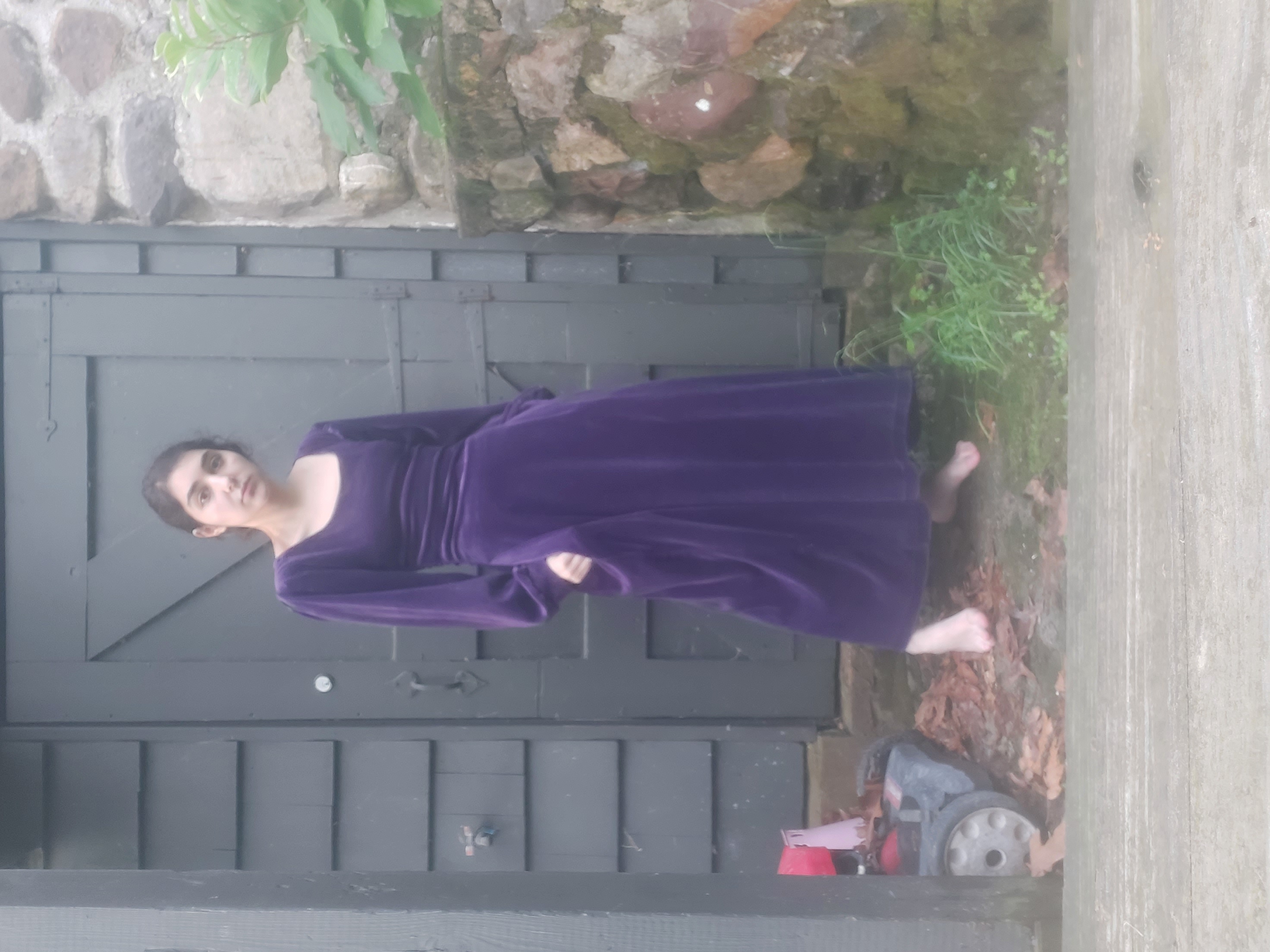 a young woman stands barefoot in a longsleeved, purple velvet maxi dress in front of a grey doorway and a stone wall. a lawnmower is visible to the left. the image appears to be an outdoor portrait, focusing on the woman and her dress.