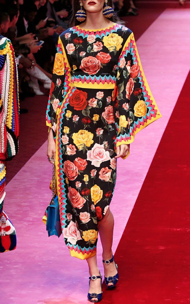 a model walks the runway at a dolce & gabbana fashion show. she is wearing a vibrant midi dress adorned with a large floral print featuring red, yellow, and pink roses on a black background. the dress has long, wide sleeves with a colorful scalloped trim, and a fitted bodice.  she is accessorized with large statement earrings and a blue handbag, and she's wearing highheeled shoes with blue embellishments. the runway is a combination of pink and red carpeting.  the background includes other models and spectators. the overall image is bright, colorful and showcases the brand's signature bold style.