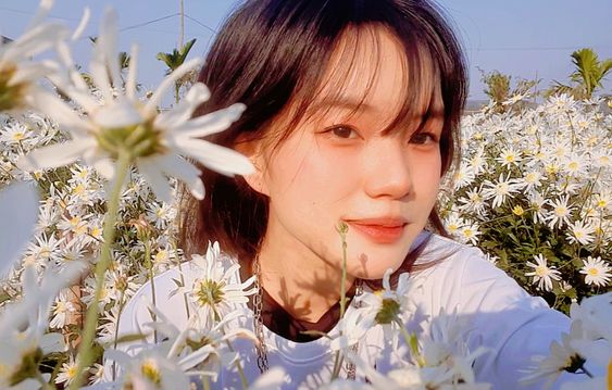 a young woman with short dark hair takes a selfie in a field of white daisies. she is wearing a white shirt and a silver chain. the background is blurry and the lighting is bright, highlighting the woman's face and the white flowers.