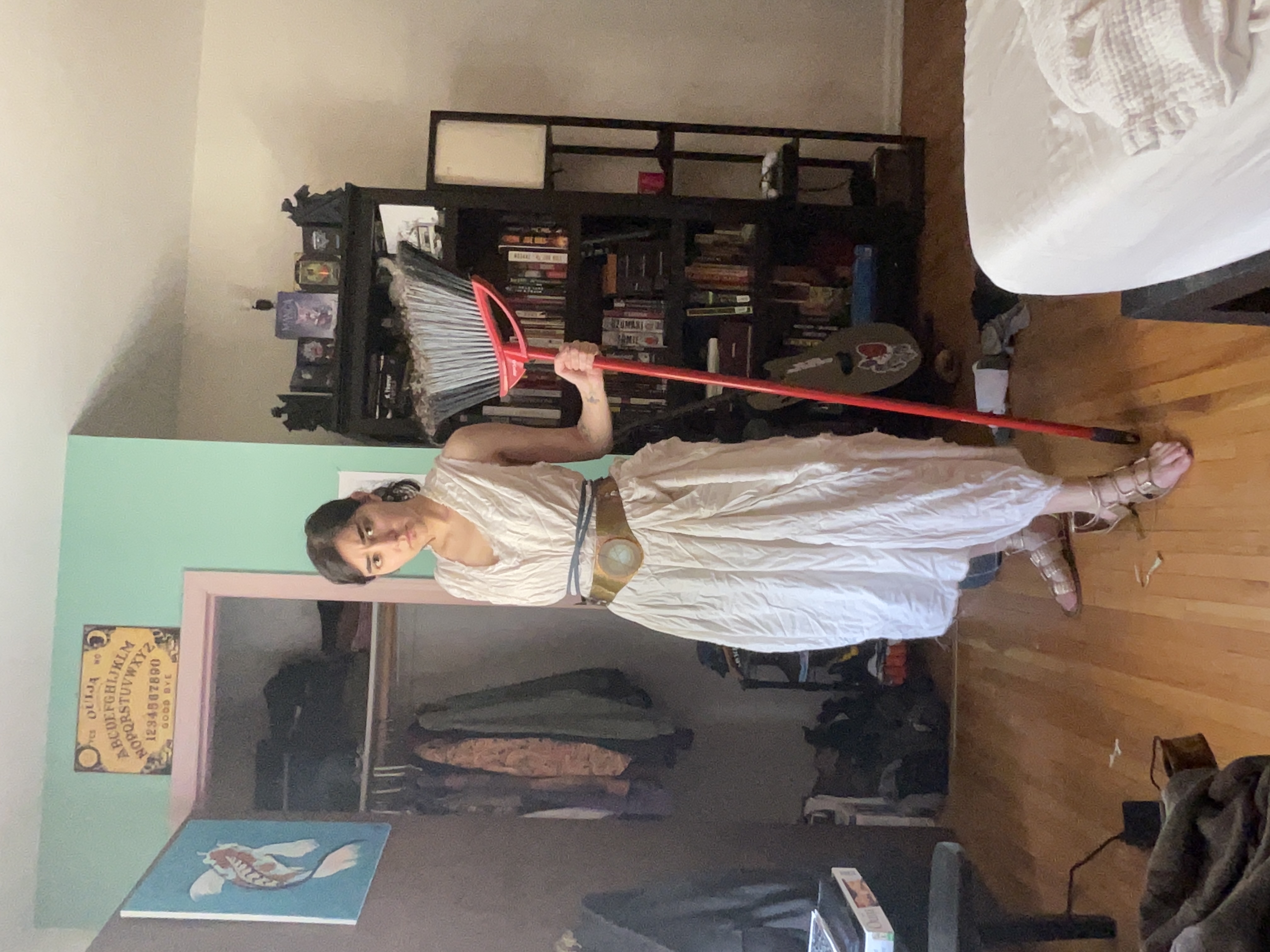 a woman stands indoors holding a red broom. she is wearing a white dress with a brown belt and sandals. behind her is a bedroom with a closet, bookshelf, guitar, and bed. above the closet is an ouija board.