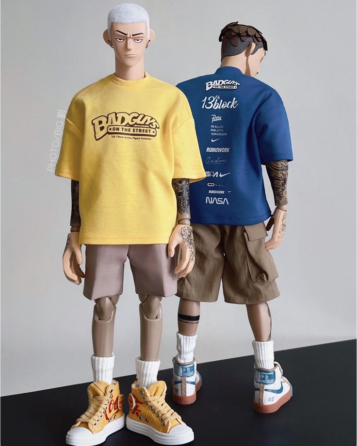the image shows two male figures standing side by side. they are wearing tshirts and shorts. the figure on the left is wearing a yellow tshirt with a "badguys" logo and khaki shorts. the figure on the right is wearing a blue tshirt with various logos and khaki shorts. both are wearing sneakers. the figures appear to be dolls or figurines.