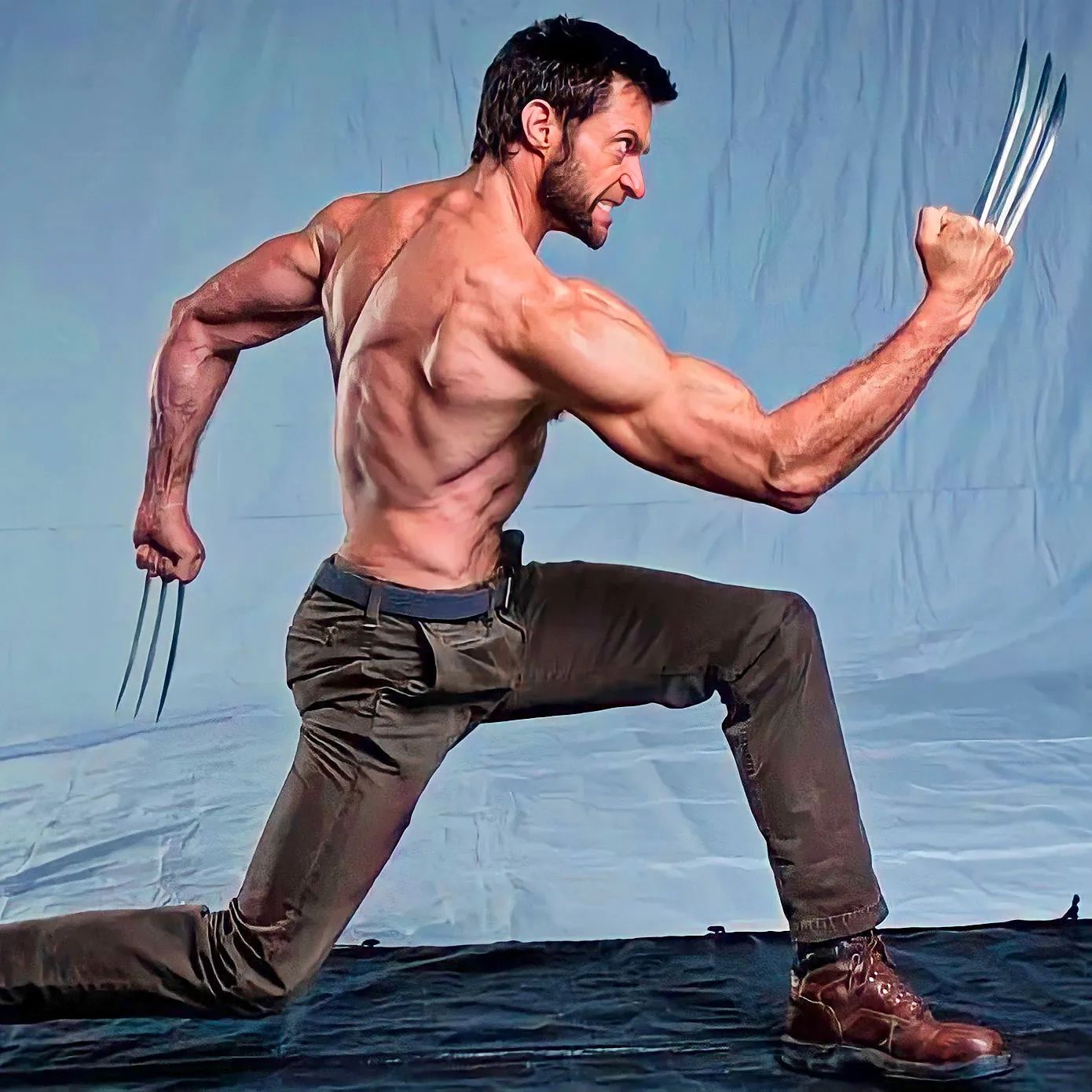 a muscular man, identified as hugh jackman portraying wolverine, is captured midaction pose against a light blue studio backdrop.  he is shirtless, revealing a toned physique, and wearing brown cargo pants and boots. his expression is fierce as he brandishes his signature adamantium claws. the lighting is soft and diffused, highlighting his muscular definition. the low angle perspective emphasizes his powerful stance.