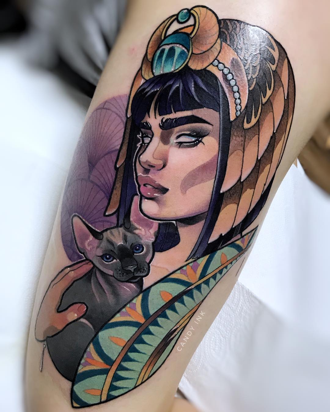 the image showcases a vibrant tattoo of a woman adorned in egyptianinspired attire, cradling a sphynx cat. the woman, depicted with a serene expression, wears an elaborate headdress reminiscent of ancient egyptian royalty, complete with a golden cobra and intricate detailing. her dark hair and the cat's dark grey fur create a harmonious contrast against her skin and the colorful background. the tattoo artist's skillful use of shading and color gradients brings depth and realism to the piece.  the artwork is signed "candy ink" discreetly below the main design.
