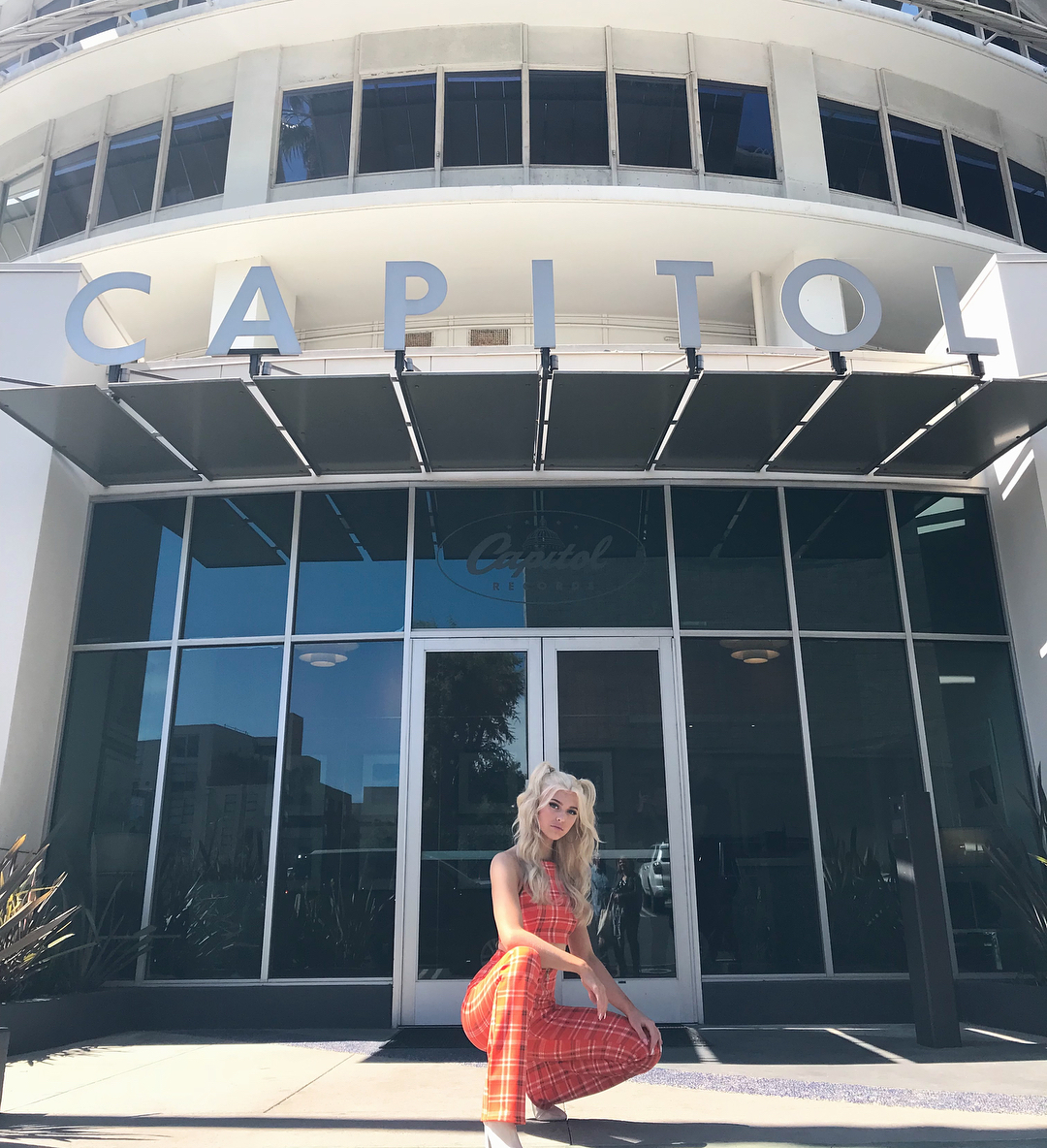 a woman with blonde hair wearing an orange plaid jumpsuit is posing in front of the capitol records building in los angeles.