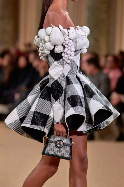 a woman model wearing a black and white checkered dress with a white florallike embellishment around the shoulder area. she is also carrying a black sequined clutch bag with a gold bletter accent. she is walking on a runway.