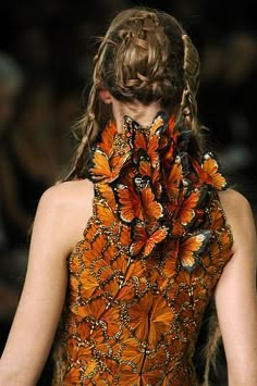 a model with long brown hair wears a beautiful orange and brown dress made of butterfly wings. the model's hair is styled in a complex braided updo and the dress is a stunning creation of intricate detail. the model is posing in a way that showcases the dress and hair beautifully, while also creating a sense of movement.