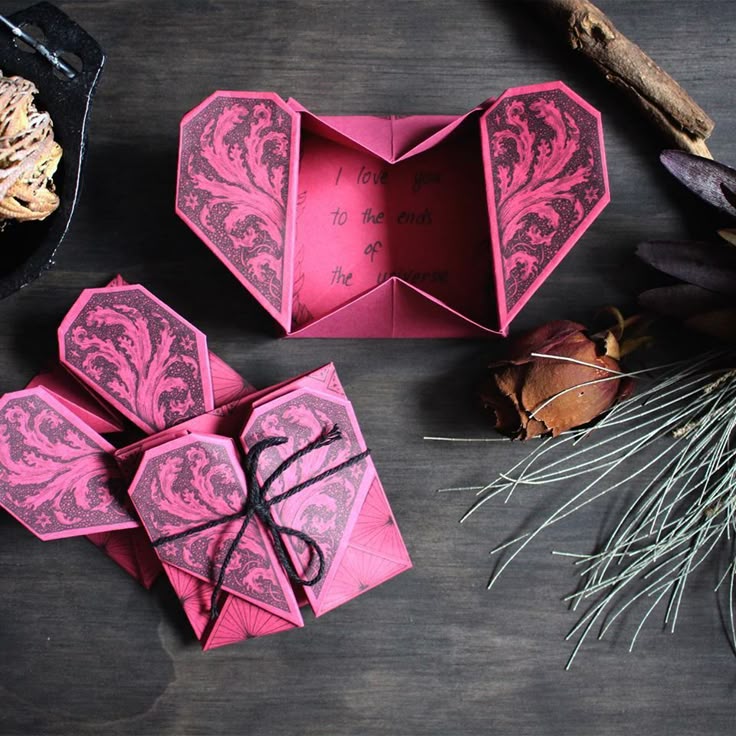 a topdown shot showcases two handcrafted origami heart boxes on a dark wooden desk. one is open, revealing a handwritten message inside stating "i love you to the ends of the universe." the other box is closed and tied with a thin black cord. both boxes are made of pink paper with a detailed dark, botanical, floral pattern on the heartshaped flaps. a withered rose, dried plants, and other decorative elements are scattered around the boxes.