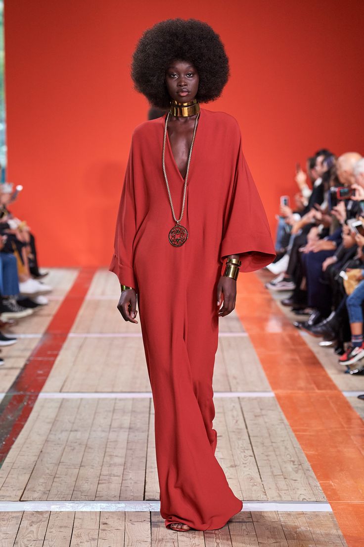 a black female model with a large afro hairstyle is showcasing a long, flowing red maxi dress. she's adorned with gold jewelry, including a choker necklace and wide bracelets.  the dress has wide sleeves and a relaxed fit.  the model walks down a runway with a wooden floor and an orange stripe running down the center. in the background, an audience is visible, watching the fashion show.  the overall setting suggests a highfashion event or runway presentation.