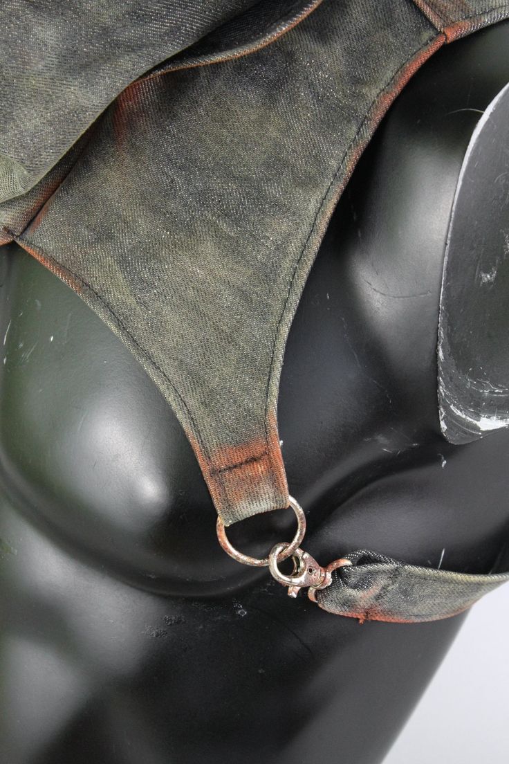 a closeup shot showcases the back detail of an olivegreen garment on a mannequin.  the fabric has a subtle tiedye effect, transitioning to a rusty orange hue towards the bottom strap. this strap features two metal rings and a clasp for adjustable fitting. the image highlights the texture and design elements of the clothing.