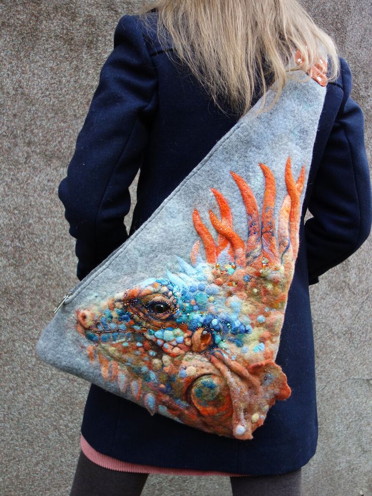 a woman wearing a navy blue coat is holding a unique and beautiful felt bag. the bag is made of felt and features a detailed and colorful needle felted iguana. the iguana's scales, eyes, and other features are all intricately crafted. the bag is worn crossbody style and hangs down to the woman's hip. the bag is a statement piece and is perfect for someone who loves unique and handmade items.