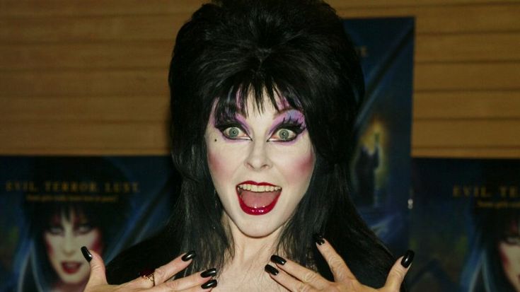 a woman with long black hair, wearing black clothing and black lipstick, is smiling at the camera. she has black nail polish on her nails. she is wearing makeup that makes her look like a witch.