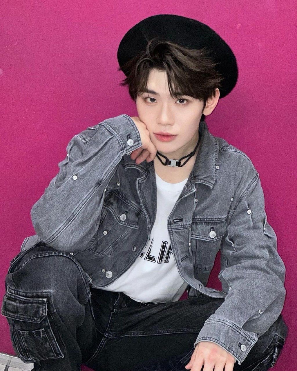 a young man with short, dark hair wearing a black beret and a denim jacket over a white tshirt.  he is posed in a squat against a hot pink wall.