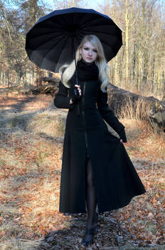 a blonde woman in a long black coat stands in a forest holding a black umbrella. the leaves on the ground are brown and orange, suggesting it is autumn or fall. she is wearing black tights and shoes and has a scarf around her neck. the overall atmosphere is gothic and elegant.