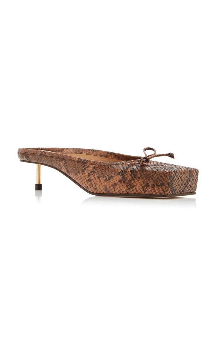 a pair of brown snakeskin mules with a low kitten heel and square toe. the shoes have a small bow detail at the top.  they appear to be amina muaddi brand, known for its stylish and luxurious designs. the image showcases the shoe from a slightly elevated angle, highlighting its features. the background is plain white, drawing all attention to the footwear.