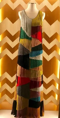 a colorful fringe dress on a mannequin against a gold chevron patterned background. the dress is made of a patchwork of colorful fringe fabric.  it is a stylish and unique piece of clothing.