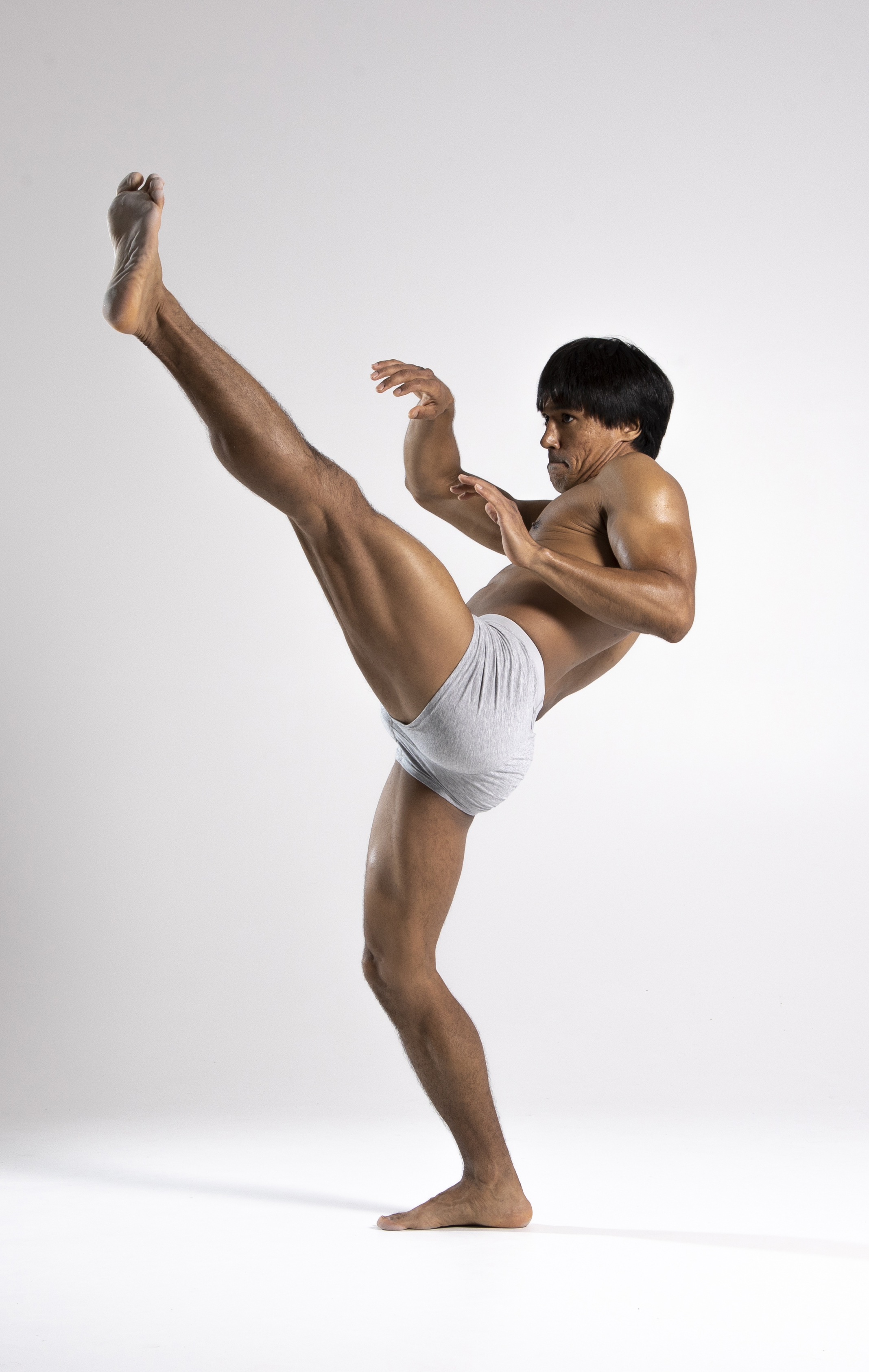 a shirtless asian man in briefs is striking a martial arts pose. he is barefoot and standing on one leg while kicking the other leg high in the air. he is in a studio with a white background.