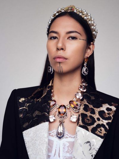 a portrait of a woman wearing elaborate jewelry and face markings. she has a pearl tiara on her head, large gemstone earrings, and a statement necklace with amber, white, and a teardropshaped jewel. she also has three vertical lines tattooed or drawn on her chin and a short horizontal line near her right temple. the woman is wearing a black blazer with white and leopard print panels. underneath the blazer, she is wearing a white lace top. she is looking directly at the camera with a neutral expression. the background is a plain light grey or white.