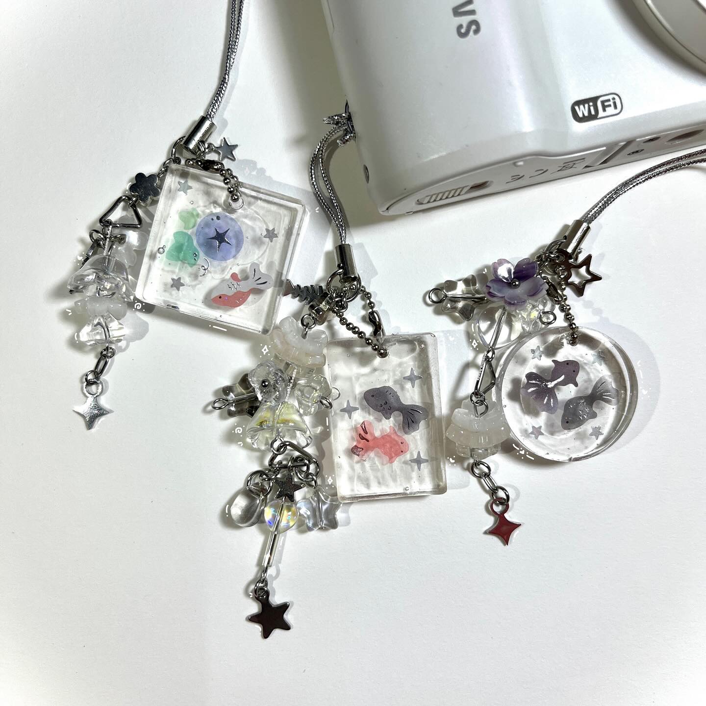 the image shows three handmade resin keychains attached to a white camera. the keychains have various shapes and feature goldfish and flower decorations, along with star charms and beads. each keychain is connected to the camera with a silver chain. they appear to be handmade jewelry or craft accessories.