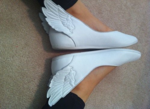 a closeup shows a pair of white ballet flats with angel wings attached to the heels. the shoes are on a person's feet, and the wings are textured to resemble feathers. the background is a beige carpet.
