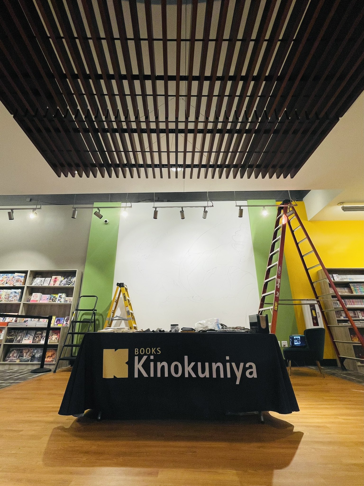 an indoor shot of a bookstore under construction. there are bookshelves lining the left and right walls, two ladders set up, and a table in the center of the room draped with a black tablecloth that reads "books kinokuniya" in large, yellow text. the ceiling has a distinctive wood slat design.