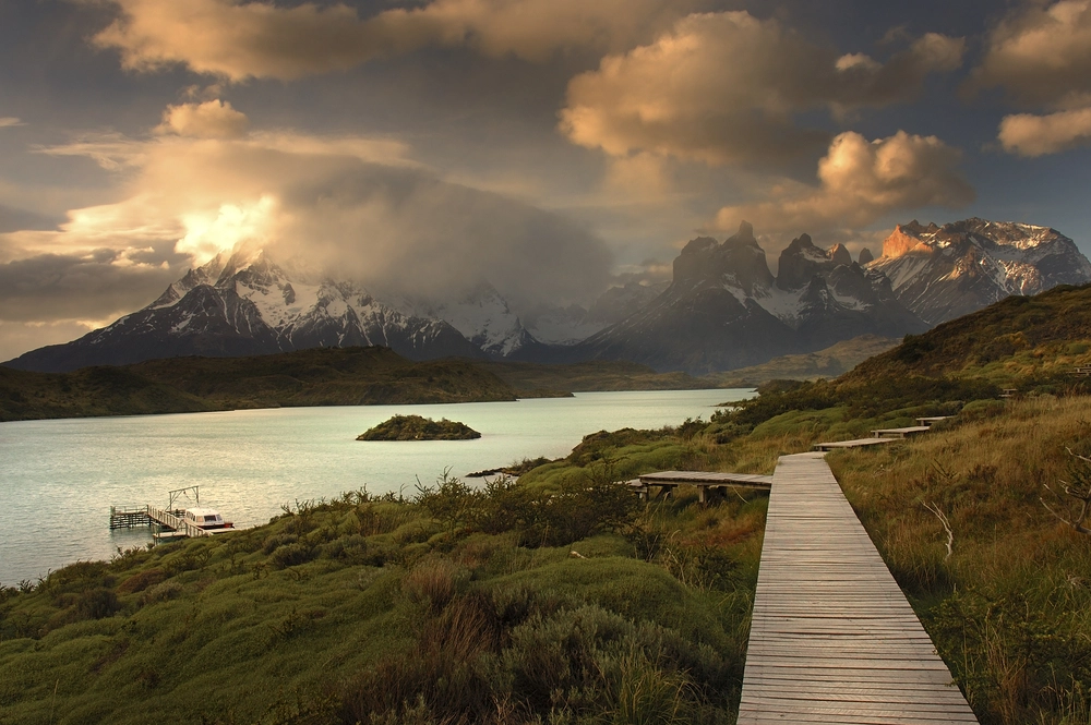 the image shows a scenic view of torres del paine national park in patagonia, chile. a wooden boardwalk leads towards a lake with a small island in the middle. in the background, there are majestic mountains, some with snowcapped peaks partially covered by clouds. a dock with a boat is visible on the shore of the lake. the sky is filled with clouds, adding to the dramatic landscape.