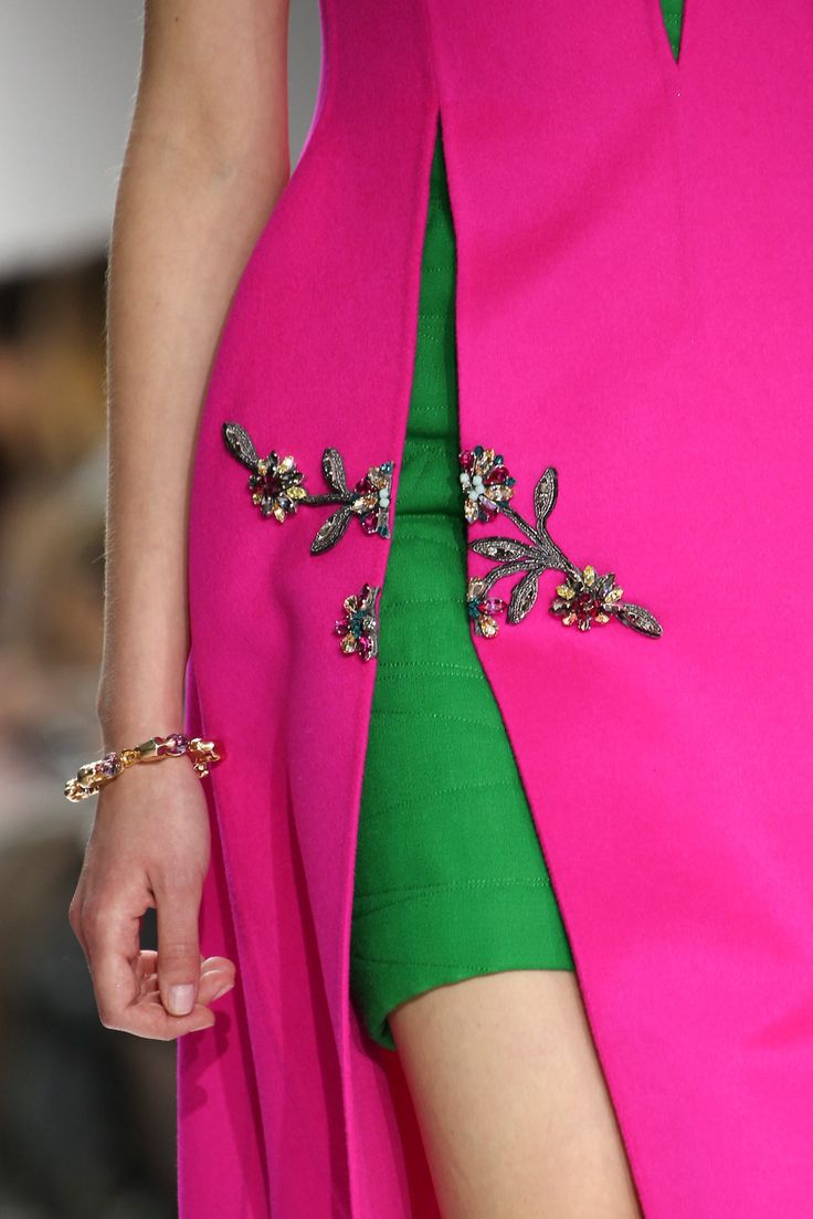 closeup view of a model showcasing a vibrant pink and green dress. the dress features a unique design with a center slit revealing a green underskirt.  intricate jeweled floral embellishments adorn the pink outer layer of the dress. the model is wearing a gold bracelet and the image focuses on the detail of the dress's construction and embellishments.  the overall impression is one of high fashion and luxurious design.