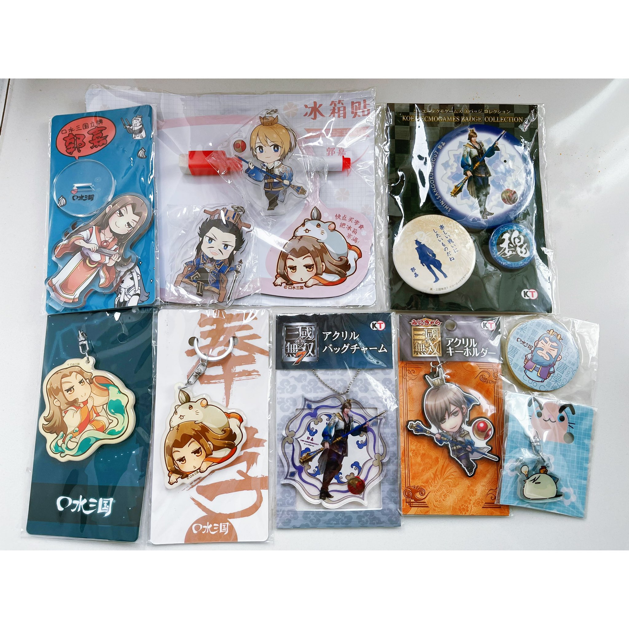 this is a collection of anime figures, charms, keychains, and buttons all featuring characters from the chinese game "three kingdoms". there are various characters from the game displayed, including one with a panda. the items are packaged and displayed in a retail setting, ready for sale.