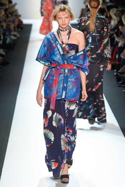 a fashion model walks the runway wearing a stylish outfit.  the ensemble is a vibrant mix of blue, red, and white floral prints.  a kimonostyle jacket is worn over a matching pair of wideleg pants.  the model has short blonde hair and is accessorized with a necklace.  another model in a similar dark floral print dress walks in the background.  the overall impression is one of high fashion and elegance.