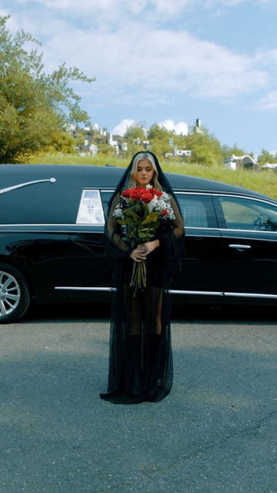 a young woman stands in front of a black hearse on a road with a cemetery and trees in the background. she is wearing a short black dress and a long, sheer black veil that reaches the ground. she holds a bouquet of red and white flowers. the sky is blue with some clouds.
