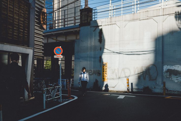 a photograph of a city street scene in japan. two men walk down a narrow alleyway between buildings. the alleyway is bordered by a tall concrete wall with graffiti and shadows cast by the sun. a no parking sign is visible. the overall mood is dark and mysterious.