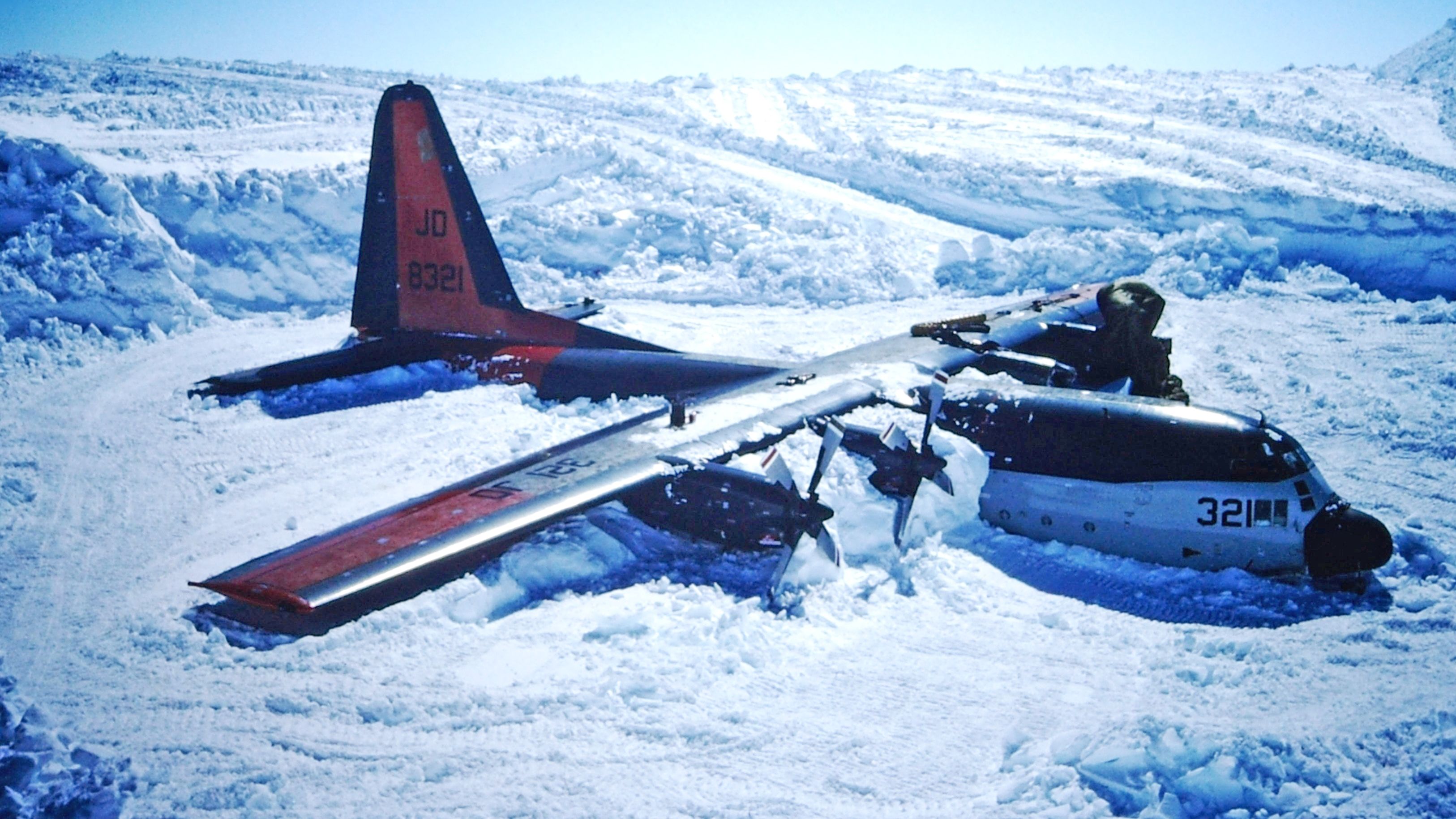 a military aircraft crashed into a snow covered field. the plane is broken in half with the rear section laying on its side and the front section laid out straight. the aircraft has a large red tail with the letters jd and the numbers 8321 on it. the front of the plane is black and white and has the number 321 on it.  there are also some snow drifts, snow banks, and ice mounds in the field.