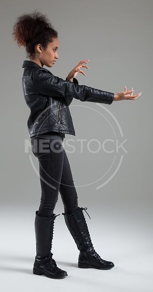 a young woman wearing a black leather jacket, jeans and boots is posing against a gray background. she is looking away from the camera and has a serious expression on her face.
