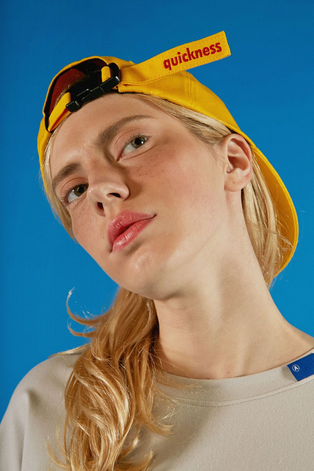 loseup portrait of a young woman with long blonde hair wearing a yellow baseball cap with the word "quickness" on the strap. she is looking off to the side and has freckles on her face. the background is a bright blue.  she is wearing a light beige, casual tshirt. the image has a clean, minimalist aesthetic.