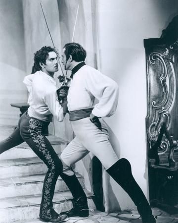 two men, dressed in period costumes, are locked in an intense sword fight. one man stands on a set of stairs, his sword held high, while the other man stands on the lower level, his sword pointing directly at his opponent. the men are both engaged in a heated battle, their expressions serious and determined. the scene is lit in a stark black and white, creating a sense of drama and suspense. the background is a shadowy, richly textured wall, with a hint of a window at the top right corner. the image evokes a sense of historical grandeur and the perilous nature of dueling.