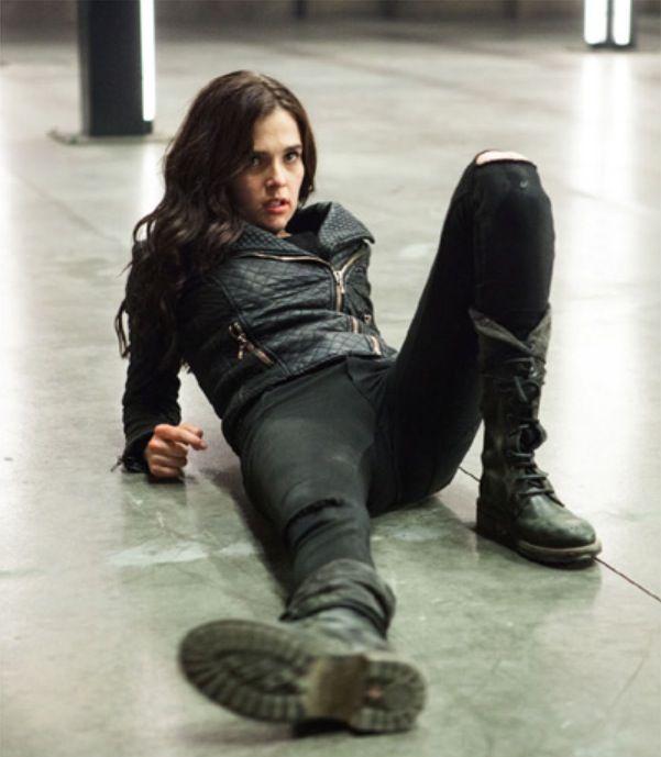 a young woman with long dark hair is wearing a black leather jacket, black jeans and boots, sitting on the floor with her knees bent and looking at the camera. she's giving a serious expression and has a cool and stylish fashion sense.