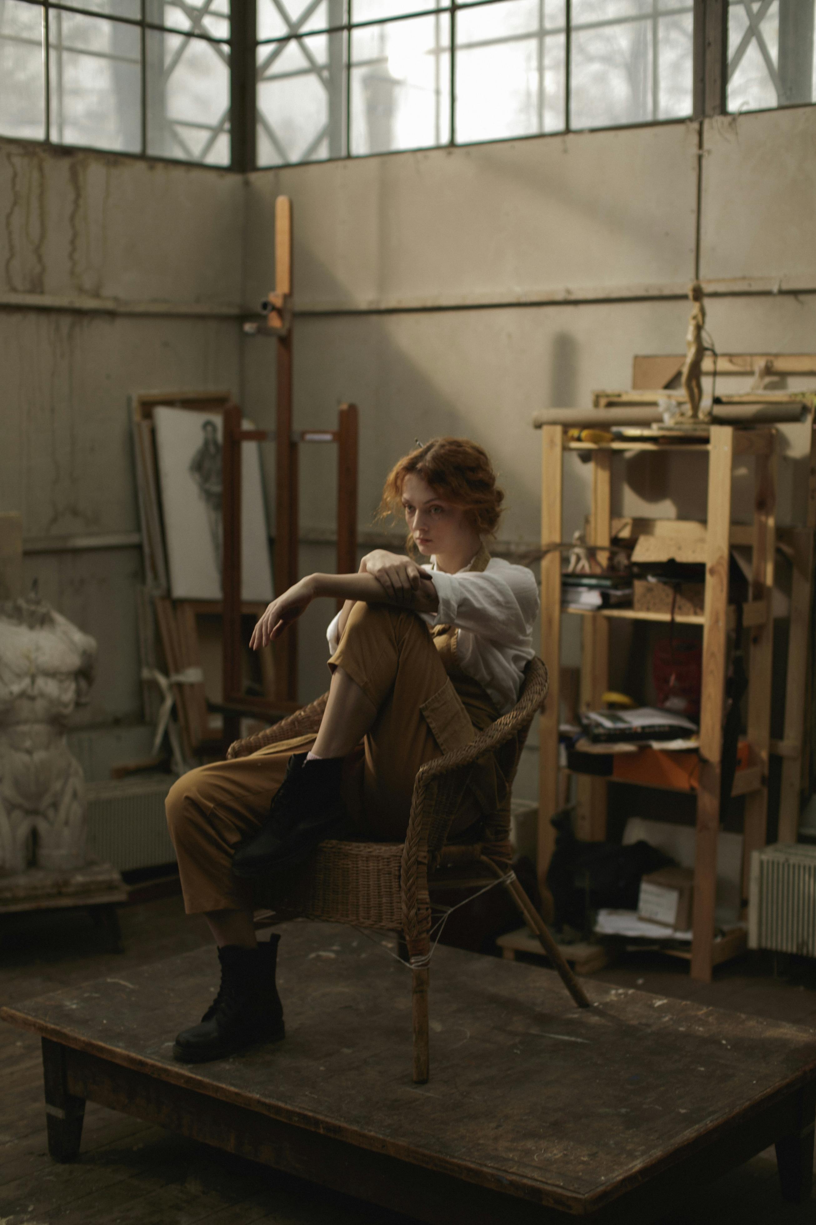 a redhead woman wearing overalls and boots sits in a wicker chair in an artist's studio. she is posed thoughtfully, with her legs crossed, and her arms folded. the studio is filled with art supplies, easels, and other paraphernalia. the overall tone is rustic and vintage. natural light streams in from large windows, illuminating the scene. the woman is the clear focal point of the image.