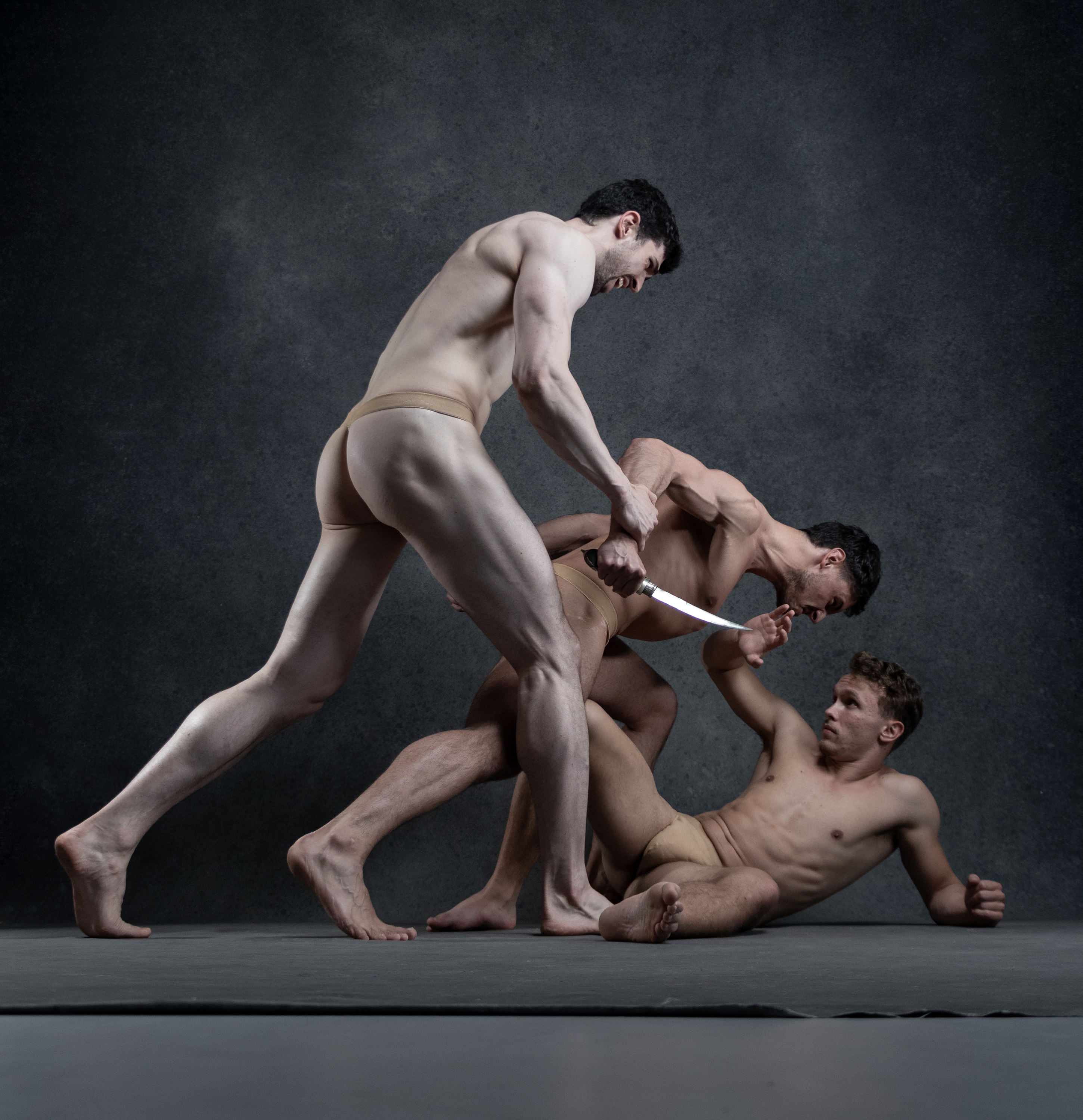 three muscular men are locked in a dynamic and tense struggle against a dark, textured background.  the scene evokes a sense of conflict and aggression, possibly a choreographed fight or dance piece. two of the men are actively engaged in a physical altercation, with one brandishing a knife, while the third appears to be caught in the middle, reacting defensively. the dramatic studio lighting emphasizes the contours of their bodies and creates a sense of theatricality.