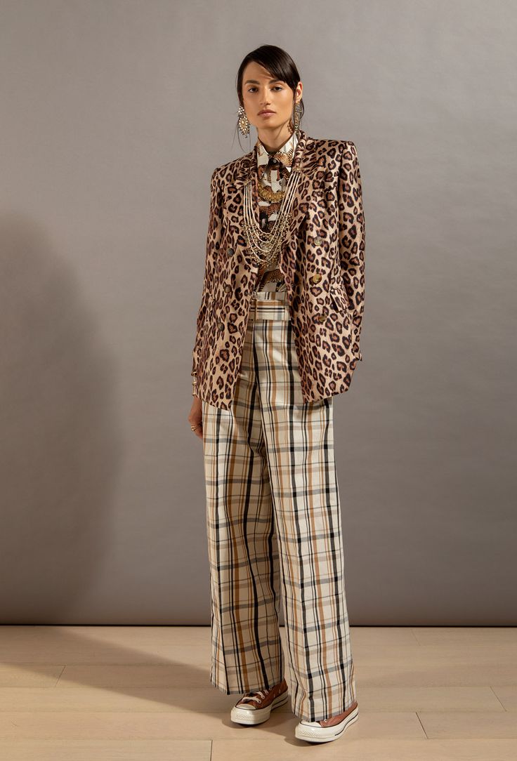 a full shot of a fashion model wearing a stylish outfit. she is wearing a leopardprint blazer over a patterned shirt, paired with wideleg plaid pants and converse sneakers.  she has on several gold necklaces and earrings. the background is a neutral beige. the overall style is a mix of casual and chic.