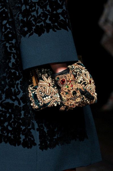 a closeup shot showcases a woman's hand, adorned with an intricately embellished clutch and gloves, emerging from a richly textured, darkgreen coat. the coat features a black velvet floral pattern. the clutch and gloves boast an opulent design, shimmering with gold threads, pearls, and colorful jewels. the overall aesthetic is one of high fashion, luxury, and rich detail, suggesting a runway or highfashion editorial image. the dark background enhances the focus on the garments and accessories.
