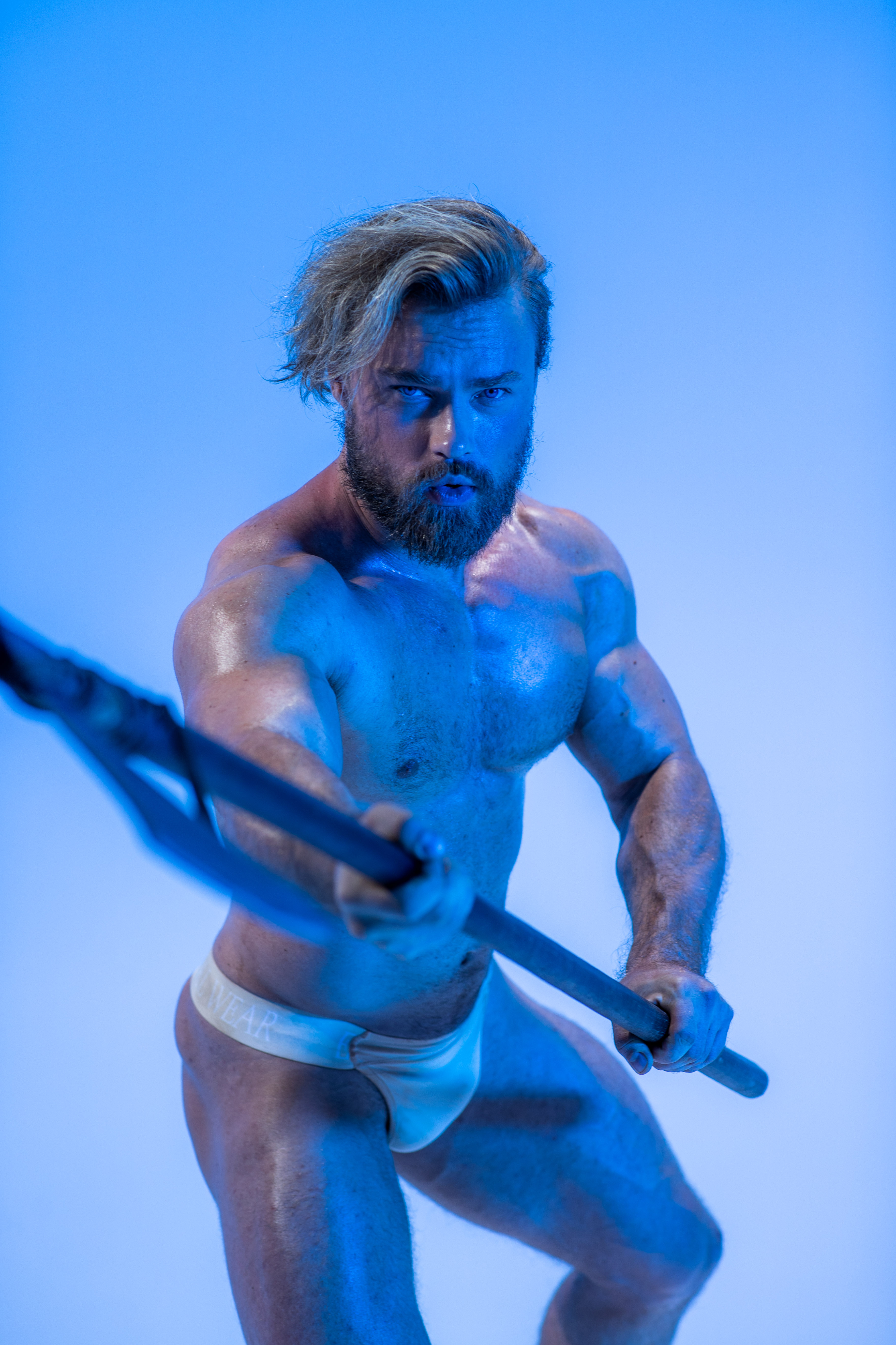 a muscular man with long blond hair and a beard stands against a vibrant blue background. he holds a black spear, his body tensed in a powerful stance. the lighting is dramatic, emphasizing his physique and creating a sense of power and intensity.