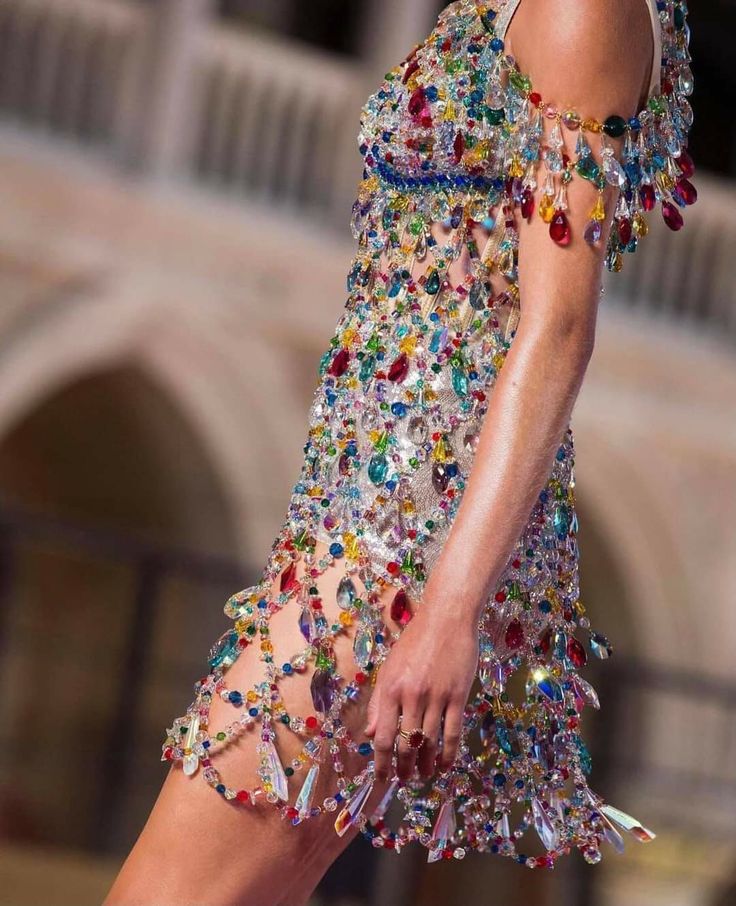 the image is a closeup shot of a model showcasing a dazzling mini dress on a fashion runway. the dress is the focal point, intricately adorned with a profusion of colorful crystals, beads, and gemstones in various shapes and sizes.  the embellishments create a shimmering, multitextured effect. the dress's design is relatively simple, but the elaborate embellishment is the defining feature. the background is blurred, drawing focus to the dress and model's arm and hand. the overall aesthetic is one of high fashion, luxury, and glamour.  the image suggests a highend fashion show or editorial photoshoot.
