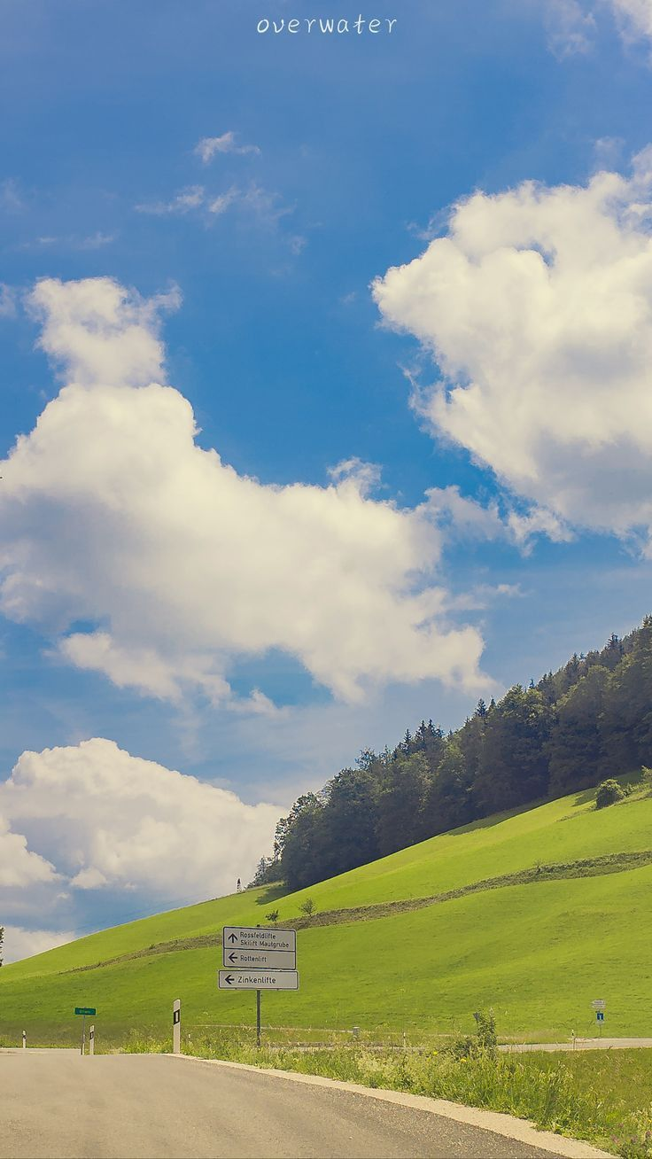 a scenic view of a green hill with trees, a road in the foreground, and a blue sky with fluffy white clouds. a road sign is visible near the road. the word "overwater" is written at the top of the image.