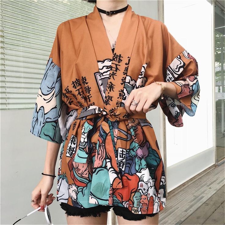 a young woman in a colorful floral kimono,  posing against a white wall with vertical blinds. she is smiling and looking at the camera. she is wearing black shorts under the kimono. she has a necklace on and is wearing a pair of sunglasses around her neck.