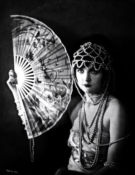 a black and white portrait of a woman in a studio setting. she is wearing a glamorous outfit and jewelry and holding a feather fan. her hair is styled in a fashionable updo and she is looking directly at the camera. the photograph has a classic hollywood feel and suggests the glamorous world of the 1920s and 1930s.
