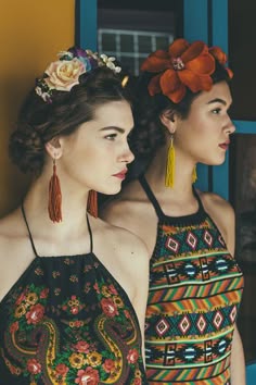 two young women are posing for a portrait. they are standing next to each other, looking towards the camera. they are both wearing colorful outfits and have flowers in their hair.  they are both wearing floral print halter tops. the women are both smiling, and they seem to be enjoying each other's company.  the background is a bright yellow wall and a blue door, creating a colorful and vibrant scene.