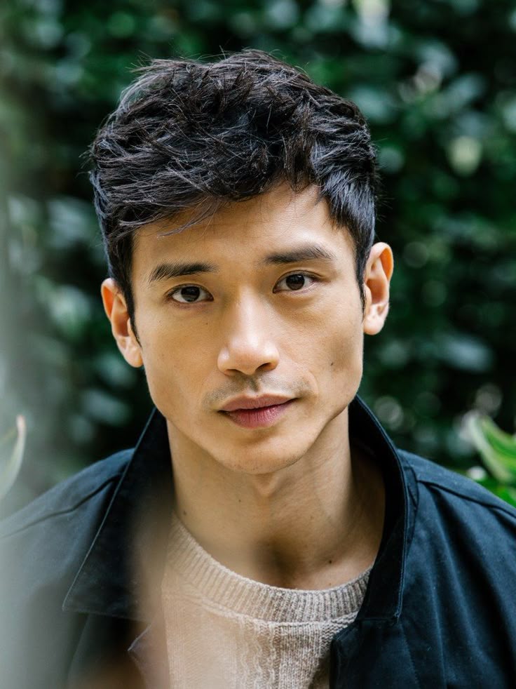 a portrait of an asian man with dark hair, wearing a jacket and sweater, looking at the camera outdoors.