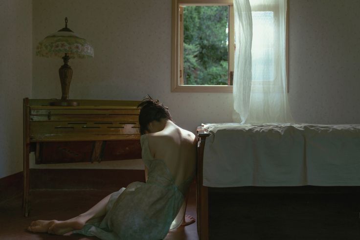 a woman in a lightcolored dress sits on the floor in a bedroom, leaning against the side of a bed. a window with a white curtain is visible, letting in natural light. a piano with a lamp on top is next to the woman. the woman's bare back is partially visible. the room has wallpaper and a wooden floor.