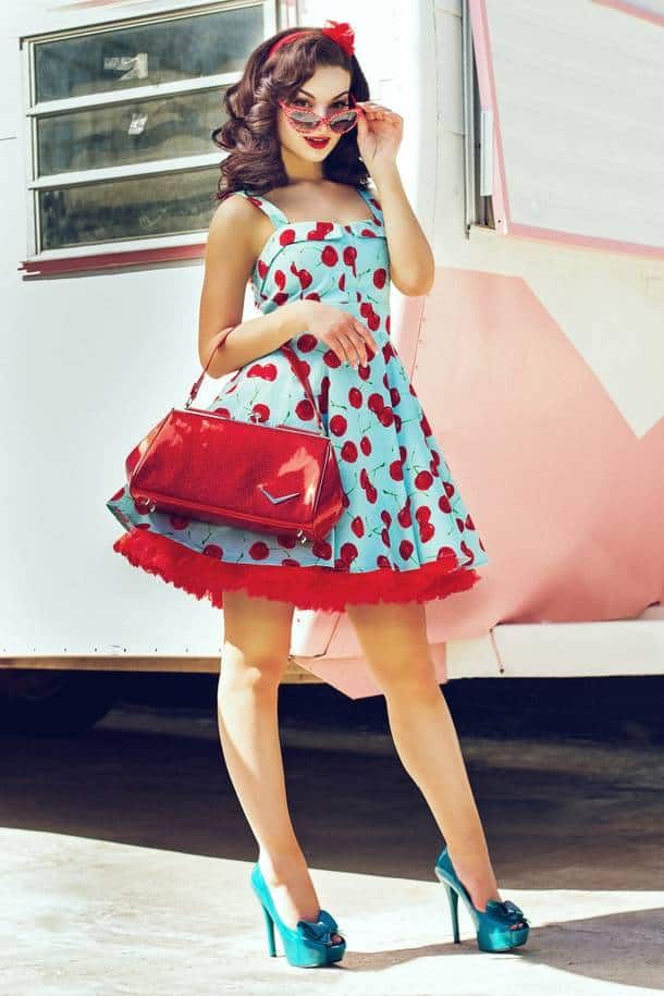 a beautiful woman in a red cherry dress and blue heels is standing in front of a retro trailer. she is wearing sunglasses and a red handbag, and her hair is styled in a vintage way. the overall feel of the image is very fashionable and glamorous.